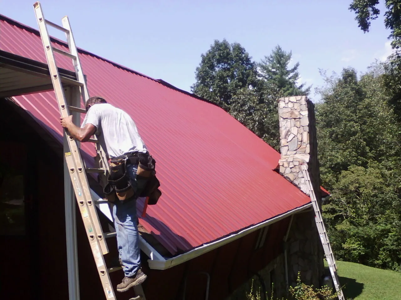 Roofer on a ladder beside a red metal roof and stone chimney — Soffit & Fascia Repair by Roofing Brothers in Wycombe